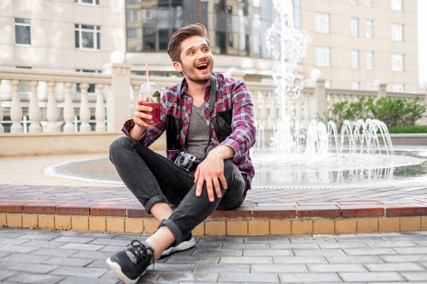 Porträt eines jungen bärtigen Mannes im karierten Hemd trinken Saft im Freien in der Innenstadt auf Brunnenhintergrund - Foto, Bild