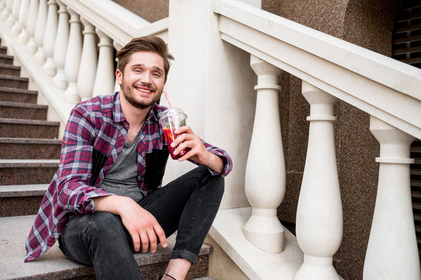 portrait of happy young bearded man in plaid shirt holding fresh juice sitting on stairs  - Photo, image