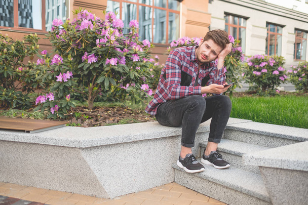 portrait of sad bearded man in plaid shirt sitting on stone stairs outdoor with pink flowers on background - Foto, imagen