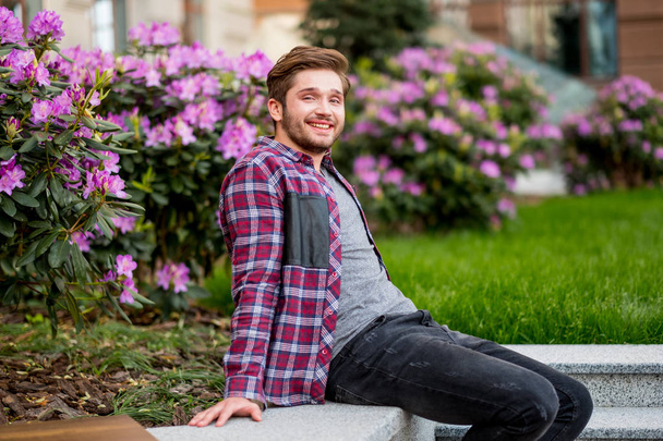 portrait of young bearded man in plaid shirt outdoor with pink flowers on background - Foto, immagini