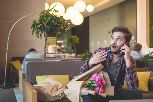 portrait of young bearded man in plaid shirt holding bouquet of flowers and using mobile indoor on grey sofa background  - 写真・画像