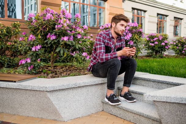 portrait of young bearded man in plaid shirt sitting on stone stairs outdoor with pink flowers on background - Photo, Image