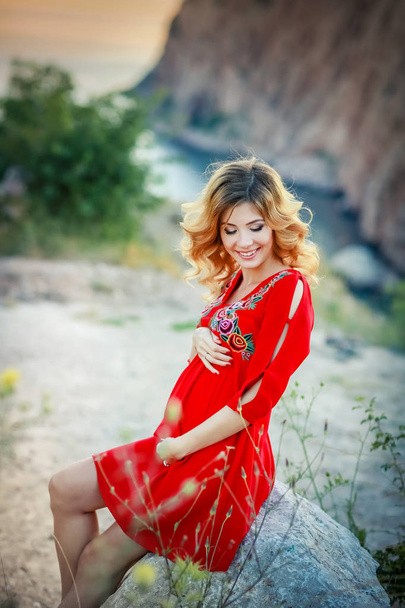 beautiful blonde pregnant woman wearing red dress posing against amazing nature landscape  - Foto, imagen