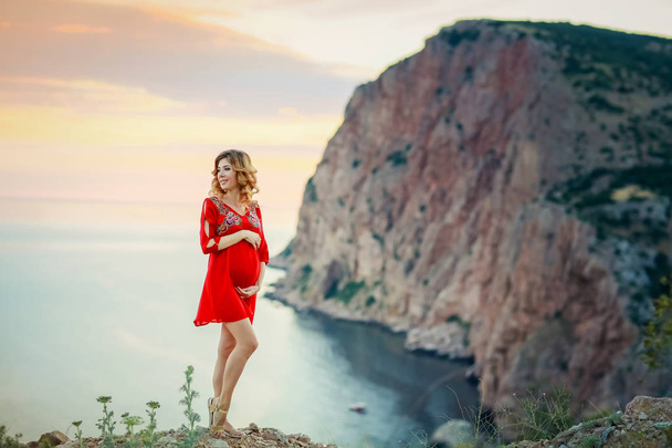 beautiful pregnant woman posing outdoors against amazing nature landscape wearing red dress  - Фото, изображение