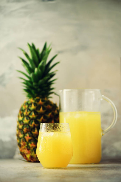 Pineapple juice in glassware and whole pineapple fruit on gray background. Copy space, sunlight effect. Summer, holiday concept. Raw, vegan, vegetarian, clean eating diet. - Φωτογραφία, εικόνα