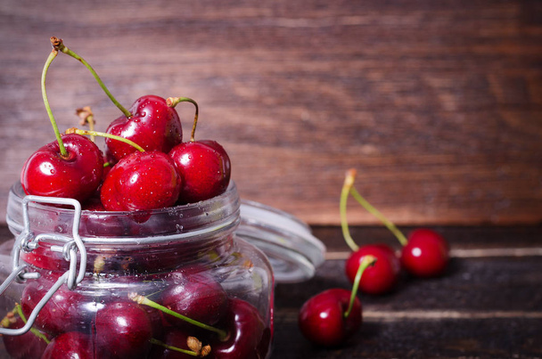 Red cherries in a glass jar on dark wooden background with copy space. Sunny summer and harvest concept. Cherry macro. Sunlight affect. Vegan, vegetarian, raw food - Photo, Image