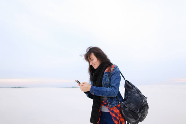 Стильная женщина в джинсах и сумке, с помощью смартфона и стоящая на белом зимнем фоне
. - Фото, изображение