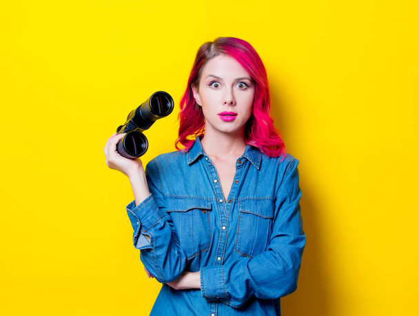 Young pink hair girl in blue shirt holding a binoculars. Portrait on isolated yellow background - 写真・画像