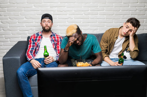 Zdenerwowany znajomych oglądać piłki nożnej z piwa i przekąski smutny z stracić ich zespołu, siedząc na kanapie - Zdjęcie, obraz