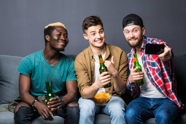 Portrait of stylish, best, attractive, modern positive guys with hairstyle, bristle sitting on couch, having mugs with lager in hands, shooting selfie on smart phone, enjoying, spending time - Foto, Imagen