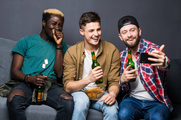 attraktive Kerle trinken Bier, machen Selfies mit dem Smartphone und lächeln, während sie zu Hause auf der Couch vor dem Fernseher sitzen - Foto, Bild