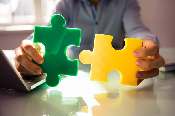 Close-up Of Businessperson's Hand Holding Two White Jigsaw Puzzle Over Desk - Photo, Image