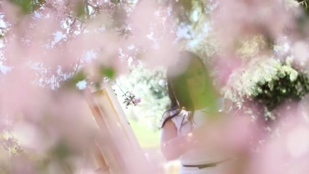 Beyaz elbiseli, sanatçı boya bir resim içinde çiçek açan çiçeklerin güzel kadın ressam bahar elma bahçesi, o bir palet boya ve bir fırça ile onun elinde tutan - Video, Çekim