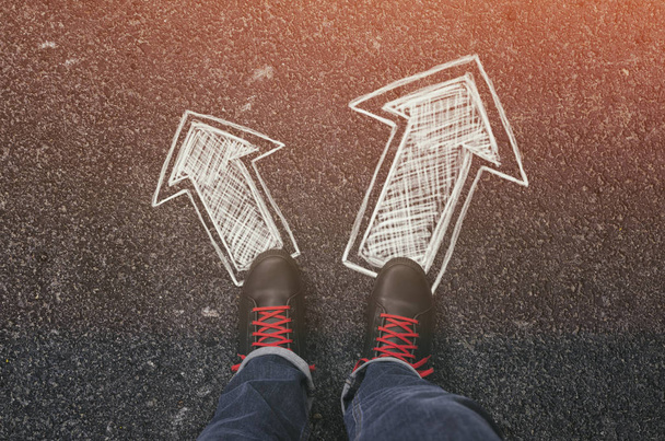 Sneakers on the asphalt road with drawn arrows pointing in two directions. Making decisions and making choices concept. - Фото, изображение