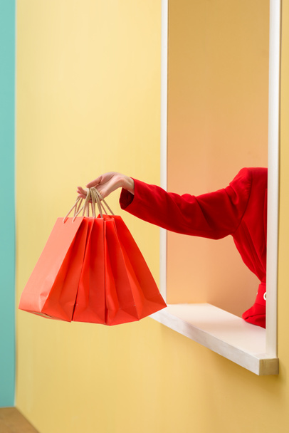 partial view of stylish woman in red clothing with red shopping bags outstretching hand out decorative window - Foto, imagen