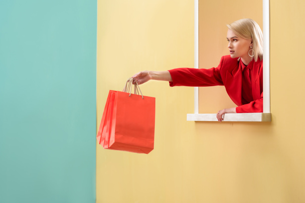 jovem mulher na moda em roupas vermelhas com sacos de compras vermelhos olhando para fora janela decorativa
 - Foto, Imagem