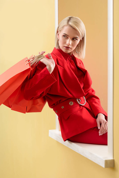young fashionable woman in red clothing with red shopping bags sitting on decorative window - Foto, afbeelding