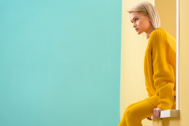 Seitenansicht einer stilvollen Frau in gelbem Pullover und Strumpfhose, die am dekorativen Fenster sitzt - Foto, Bild