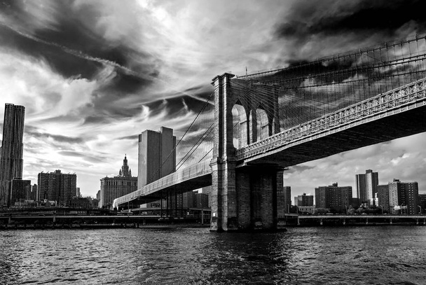 Famoso ponte di Brooklyn a New York, Stati Uniti d'America con quartiere finanziario, centro di Manhattan sullo sfondo. East River e bellissimo riflesso del tramonto. Fotografia a contrasto in bianco e nero, vista ad angolo basso
 - Foto, immagini