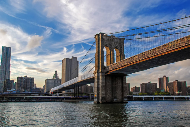 Famoso puente de Brooklyn en la ciudad de Nueva York, EE.UU. con el distrito financiero, el centro de Manhattan en segundo plano. East River y hermoso reflejo del atardecer. Vista de ángulo bajo
 - Foto, Imagen