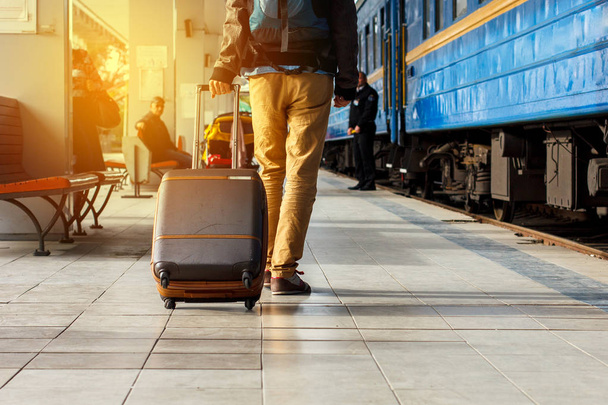 Concepto de viaje. Los hombres usan zapatillas de deporte y los pantalones amarillos viajan con equipaje marrón y mochila en la estación de tren. Día soleado
. - Foto, imagen