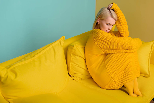 side view of young blond woman in yellow sweater and tights sitting on yellow sofa on blue backdrop - Фото, изображение