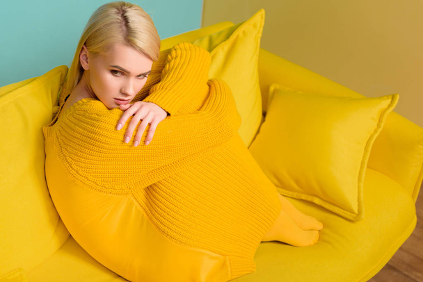 side view of young pensive woman in yellow sweater and tights sitting on yellow sofa - Fotoğraf, Görsel