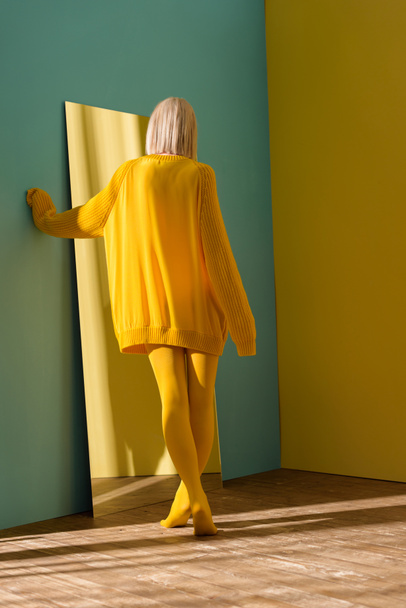 back view of blond woman in yellow sweater and tights standing at mirror - Foto, imagen