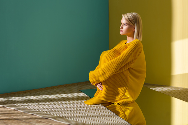 side view of pensive beautiful woman in yellow sweater and tights sitting on mirror with reflection in it - Photo, Image