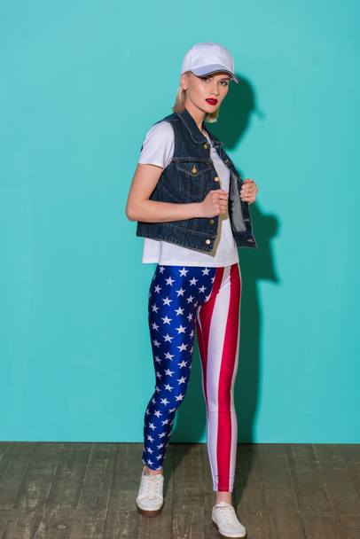 stylish young woman in cap, white shirt, denim jacket and leggings with american flag pattern posing on blue background - Fotoğraf, Görsel