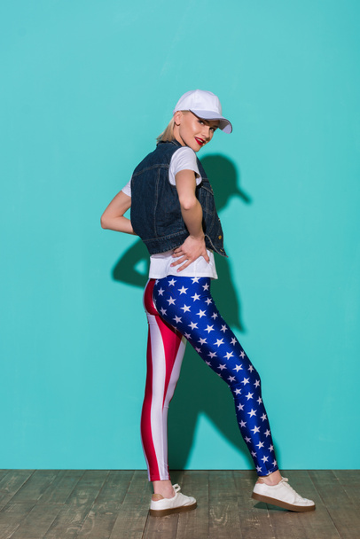 stylish young woman in cap, white shirt, denim jacket and leggings with american flag pattern posing on blue background - Photo, Image