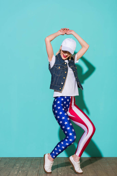 stylish young woman in cap, white shirt, denim jacket and leggings with american flag pattern posing on blue background - Photo, Image