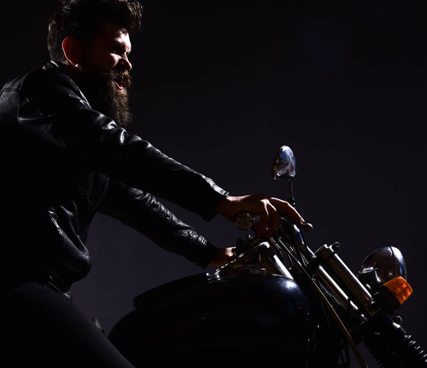 Macho, brutal biker in leather jacket riding motorcycle at night time, copy space. Masculinity concept. Man with beard, biker in leather jacket sitting on motor bike in darkness, black background - Foto, Imagem