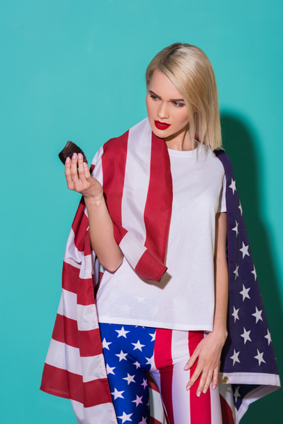 portrait of young woman with american flag and cupcake on blue backdrop, celebrating 4th july concept - Photo, Image