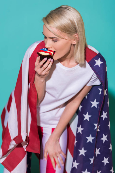 young woman with american flag eating cupcake on blue backdrop, celebrating 4th july concept - Photo, Image