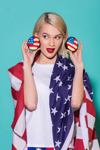 portrait of young  woman with american flag and cupcakes on blue backdrop, celebrating 4th july concept - Photo, Image