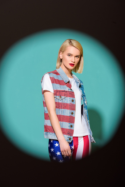 portrait of stylish young woman in denim jacket with american flag pattern posing on blue backdrop - Zdjęcie, obraz