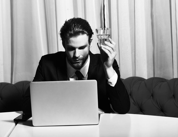 bearded man, businessman holding glass of whiskey works at laptop - Zdjęcie, obraz