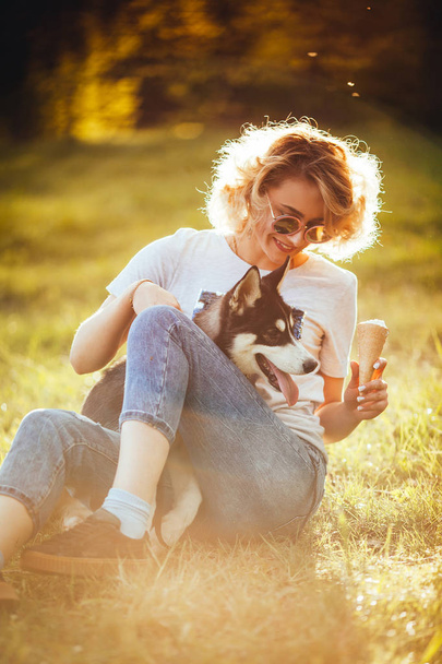 blonde with ice cream in jeans and t-shirt is sitting on the grass and holding husky in the summer in the park - Valokuva, kuva