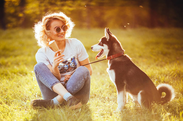 a girl in jeans and in white T-shirt with ice cream in her hand sits cross-legged in the meadow and looks at a pet in the park in the summer - Valokuva, kuva