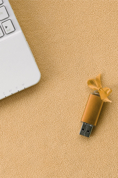 Narancs usb flash memória kártya-val egy íj fekszik egy takaró puha és Szőrős világos narancssárga gyapjú szövet mellett a fehér laptop. Klasszikus női ajándék design memóriakártya - Fotó, kép