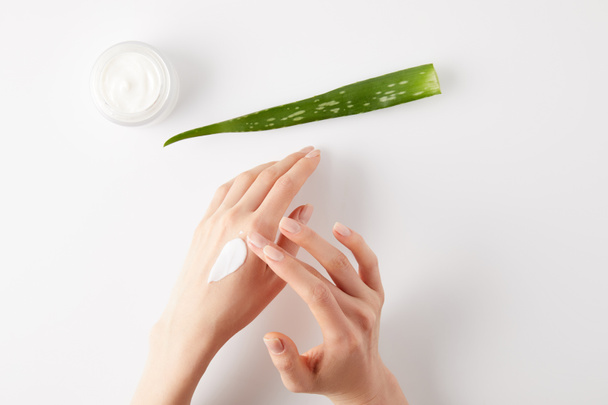 περικοπεί shot της γυναίκας που εφαρμόζουν βιολογική κρέμα για τα χέρια, aloe vera φύλλο και κρέμα εμπορευματοκιβωτίων σε λευκή επιφάνεια - Φωτογραφία, εικόνα