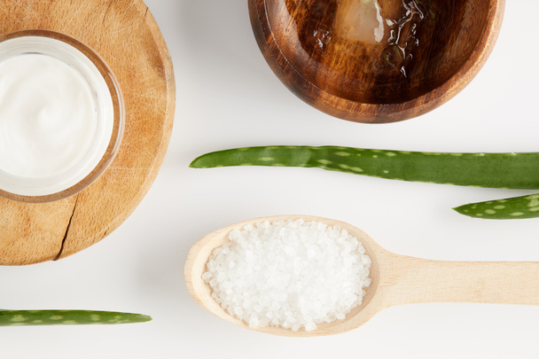 top view of organic cream in container, wooden bowl with aloe vera juice, spoon with salt and aloe vera leaves  - Fotó, kép