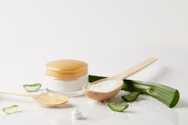 organic cream in container, spoons with aloe vera juice and salt, aloe vera leaf and slices on white surface  - Photo, image