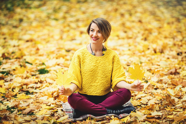 秋の草の上に座って、2 つの黄色の葉を手で保持して少女に笑みを浮かべてください。. - 写真・画像
