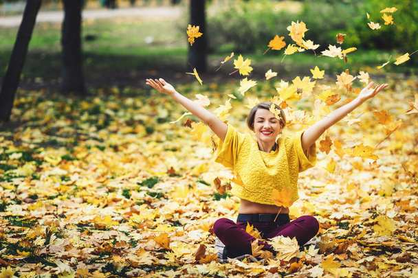 la ragazza in autunno si siede su erba e allegramente getta su foglie gialle. Stile di vita
. - Foto, immagini