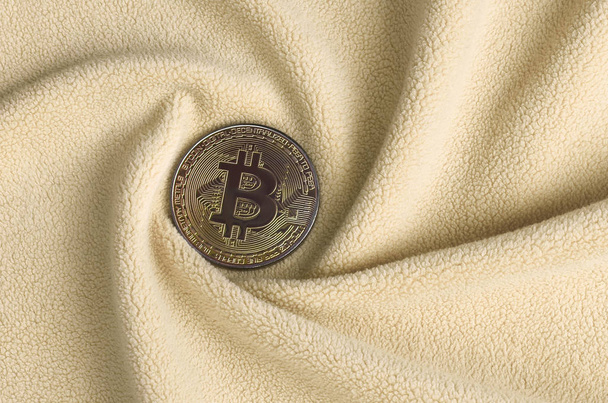 Der goldene Bitcoin liegt auf einer Decke aus weichem und flauschigem hellorangefarbenem Fleece-Stoff mit einer großen Anzahl von Relieffalten. die Form der Falten erinnert an einen Ventilator aus einem Grafikkartenkühler - Foto, Bild