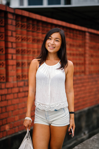 Portret van een jonge, aantrekkelijke Singaporees Chinees Aziatische vrouw met telefoon staande op een straat tegen een bakstenen muur. Ze lacht en is petite, atletische en gebruinde. - Foto, afbeelding