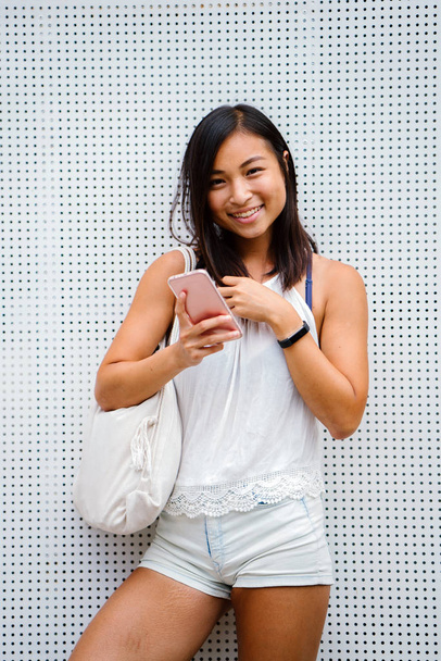 candid portrait of a cute, athletic and tanned Chinese Asian woman using her smartphone in the day on a street. She is texting and messaging her friend while she waits for someone. - Fotografie, Obrázek