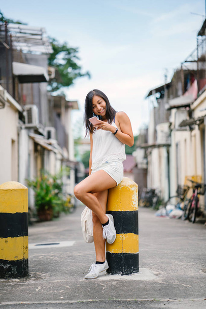 Portrait of a young, athletic, attractive and cute Chinese Asian girl using her smartphone on a street in Asia during the day. She is tanned, petite and relaxed. - Photo, image